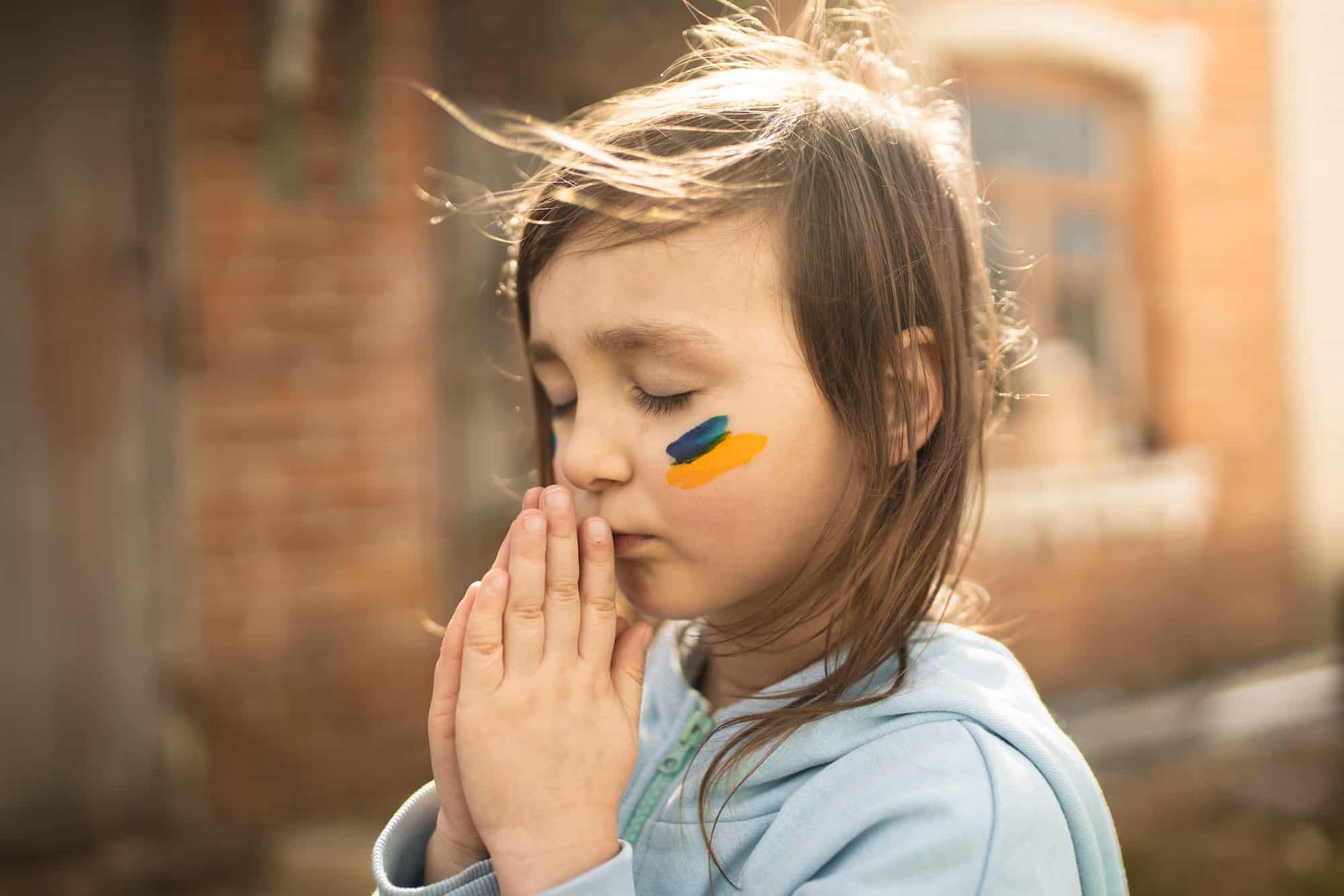 A girl with Ukrainian symbols on her face prays. A child from Ukraine. Ukrainian. Pray for Ukraine.