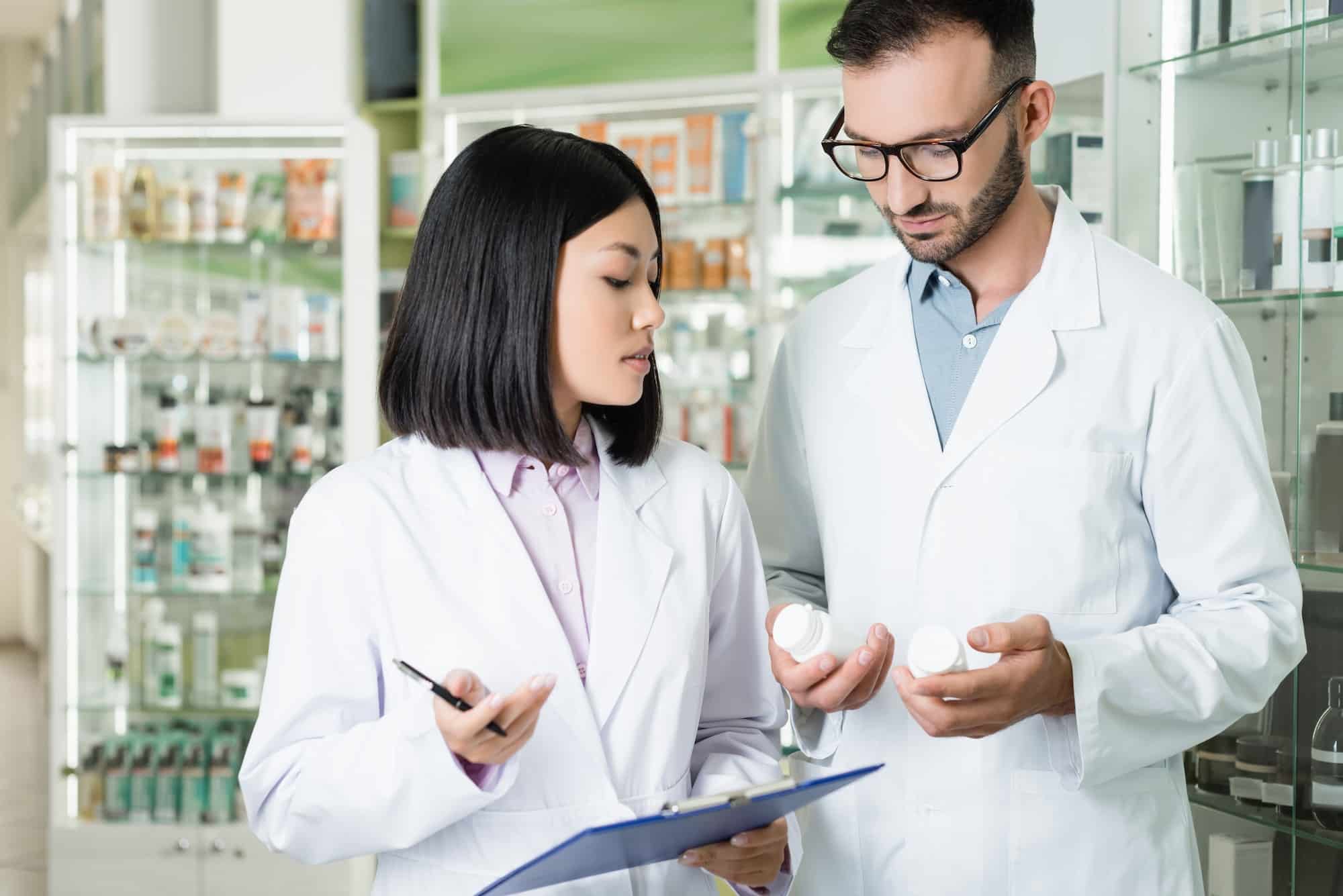 interracial pharmacists in white coats looking at bottles with medication in drugstore