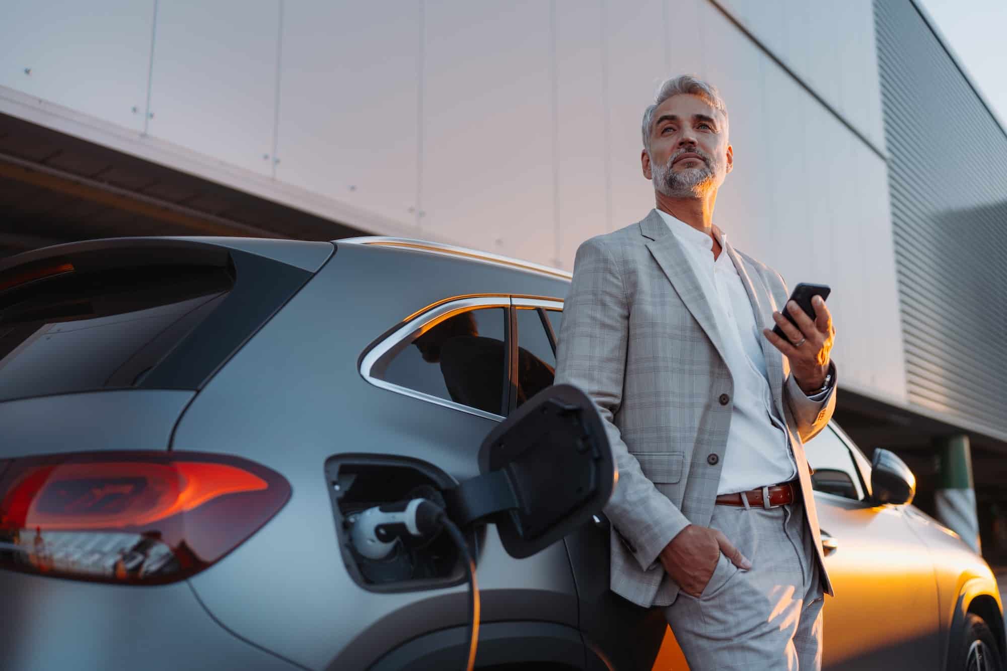 Businessman holding smartphone while charging car at electric vehicle charging station, closeup.