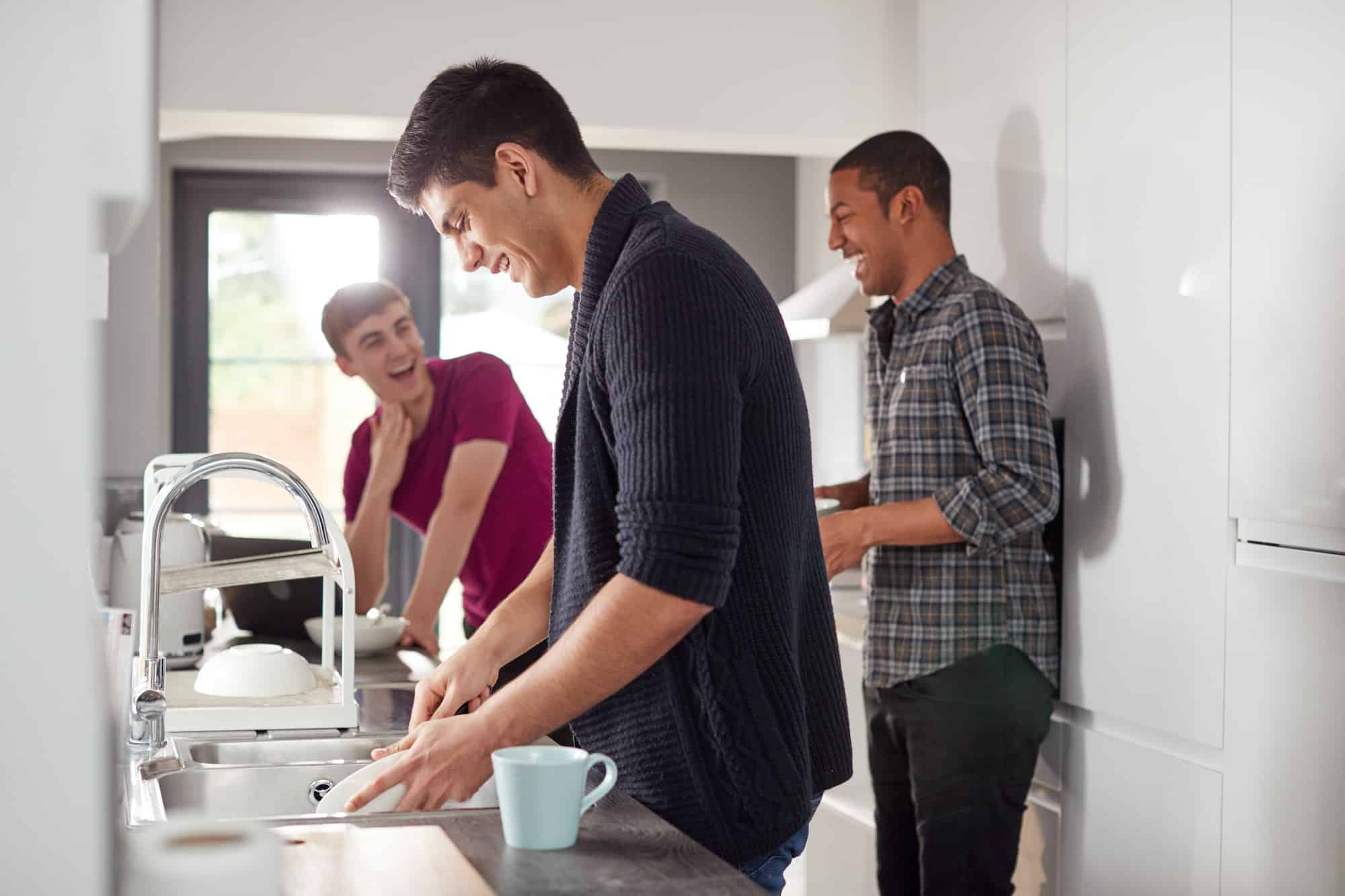 Group Of Male College Students In Shared House Kitchen Washing Up And Hanging Out Together