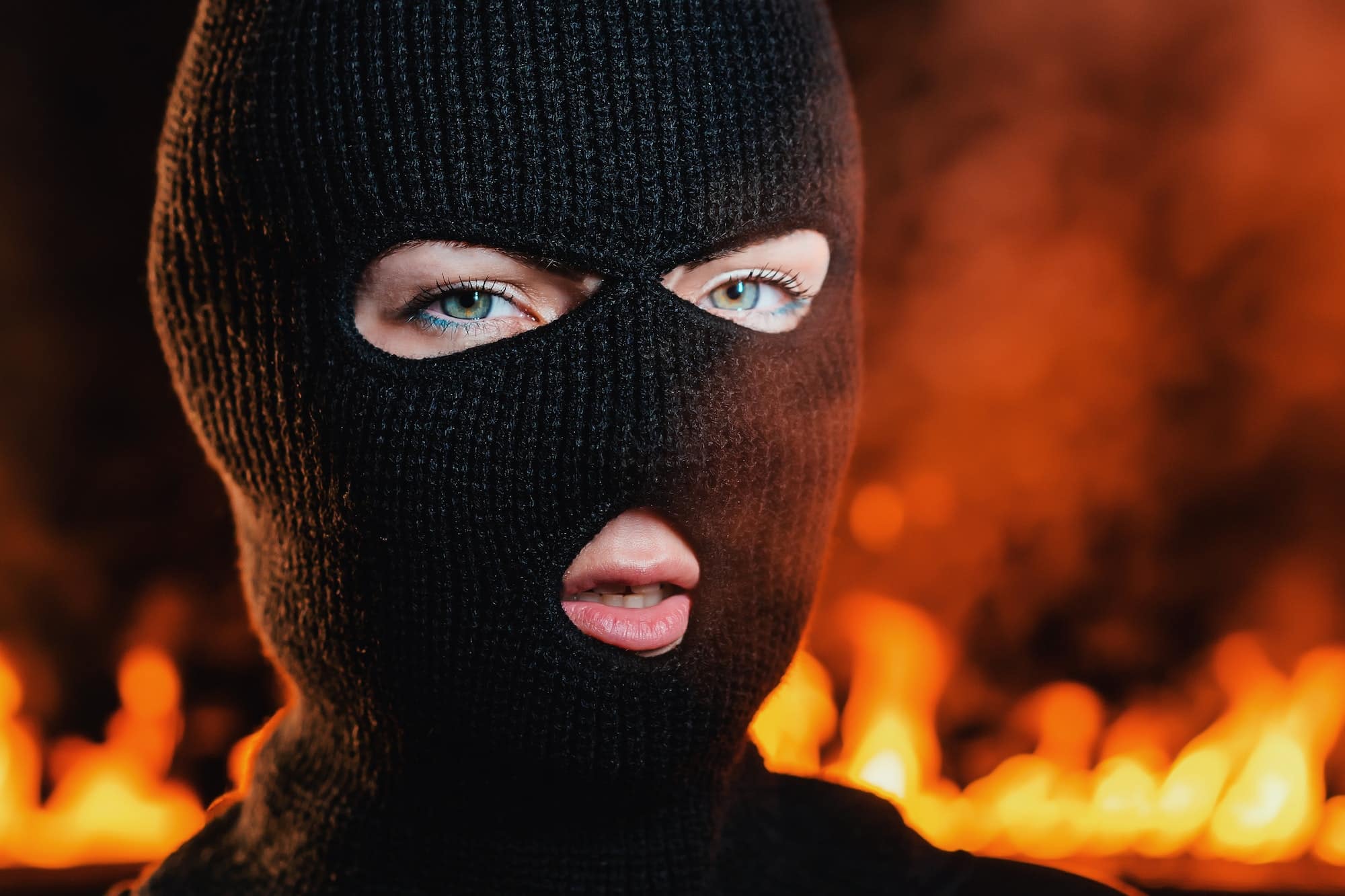 Portrait of young woman in black balaclava against backdrop of a blazing night fire.