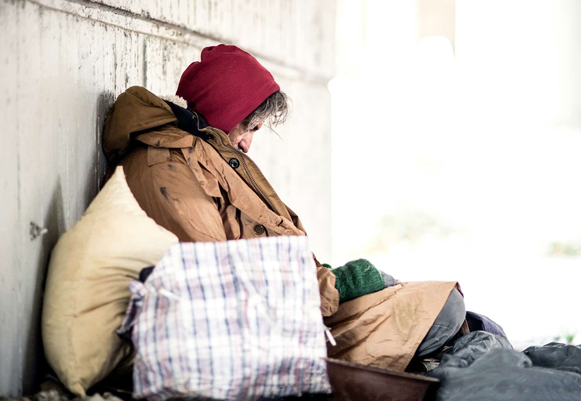 A side view of homeless beggar man sitting outdoors, leaning against a wall. Copy space.